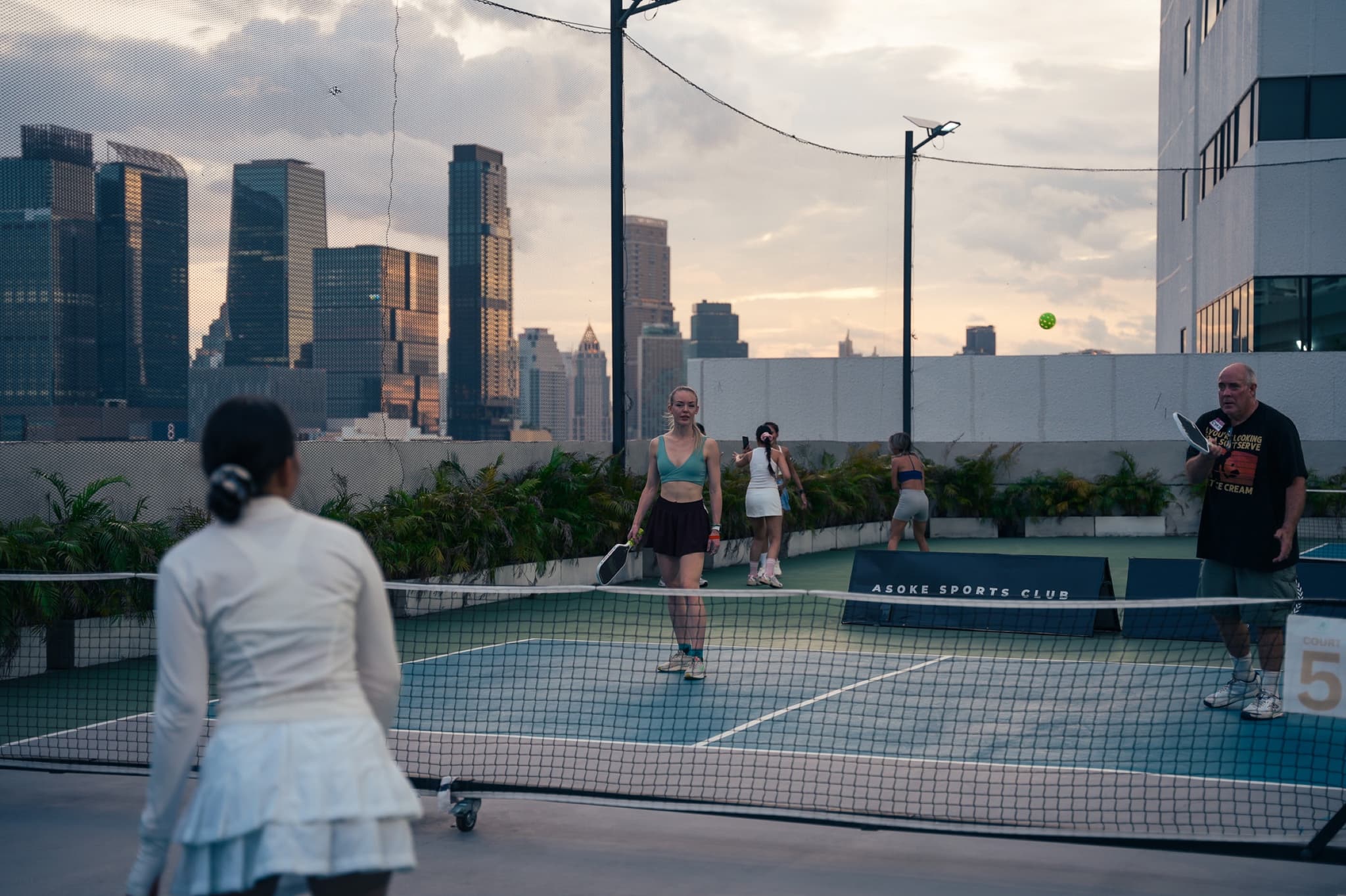 Pickleball on the Asoke Sports Club rooftop at dusk, with the Bangkok skyline behind the court