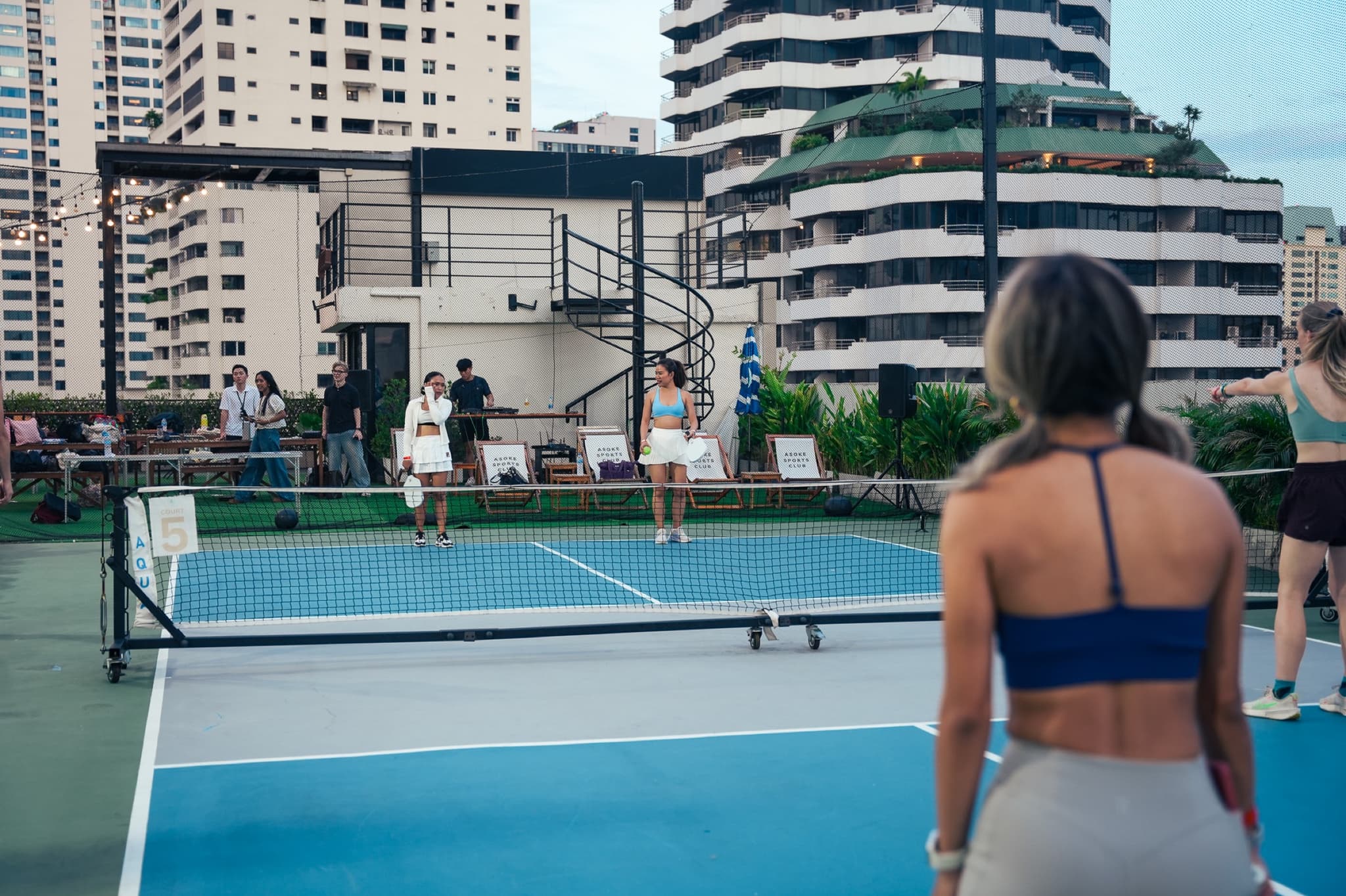 Players on a rooftop pickleball court with a DJ setup and apartment towers behind the net