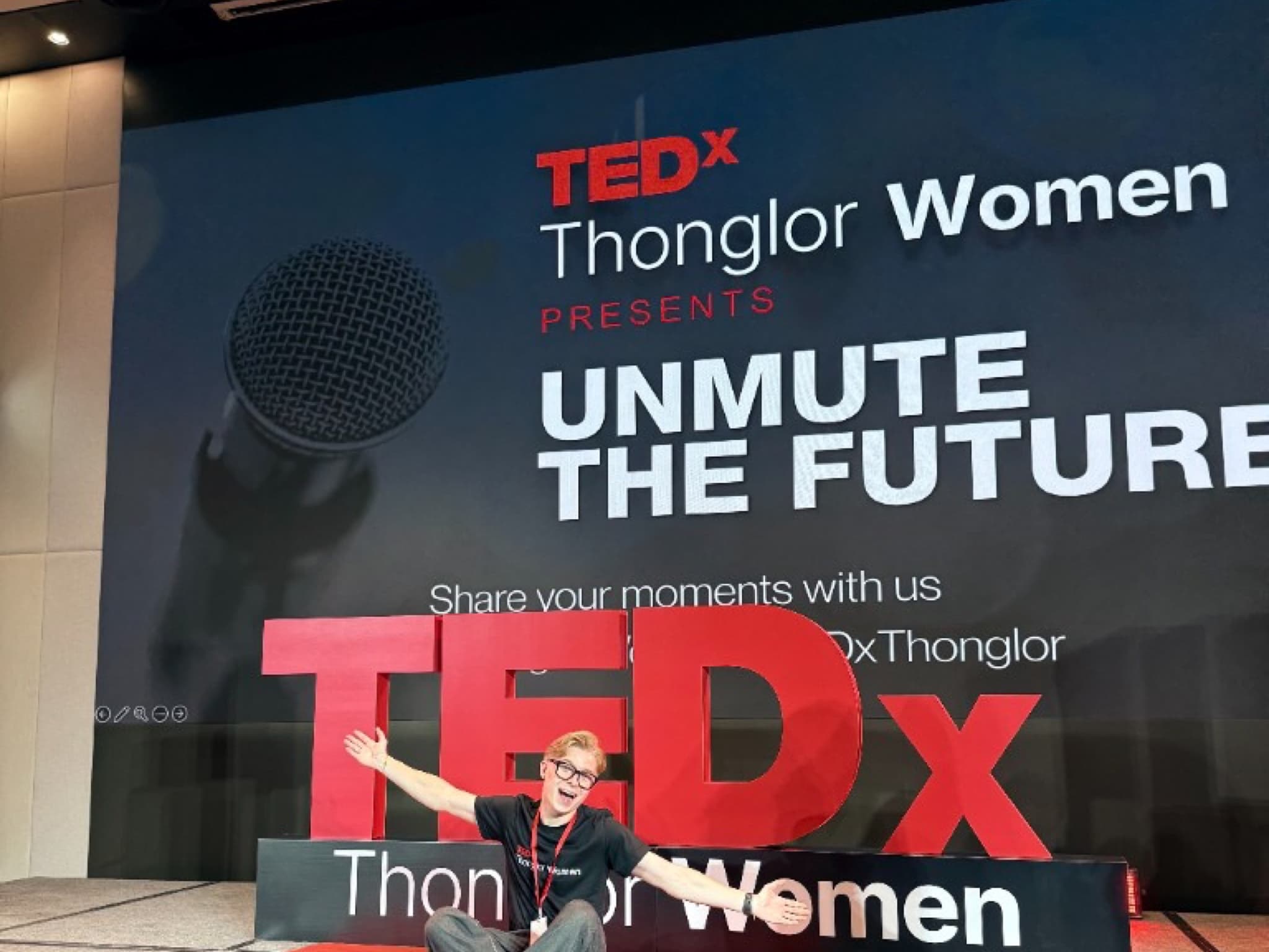 Andrei Covalescu sitting on a red rug on the TEDxThonglorWomen stage in front of the Unmute the Future backdrop and TEDx floor sign